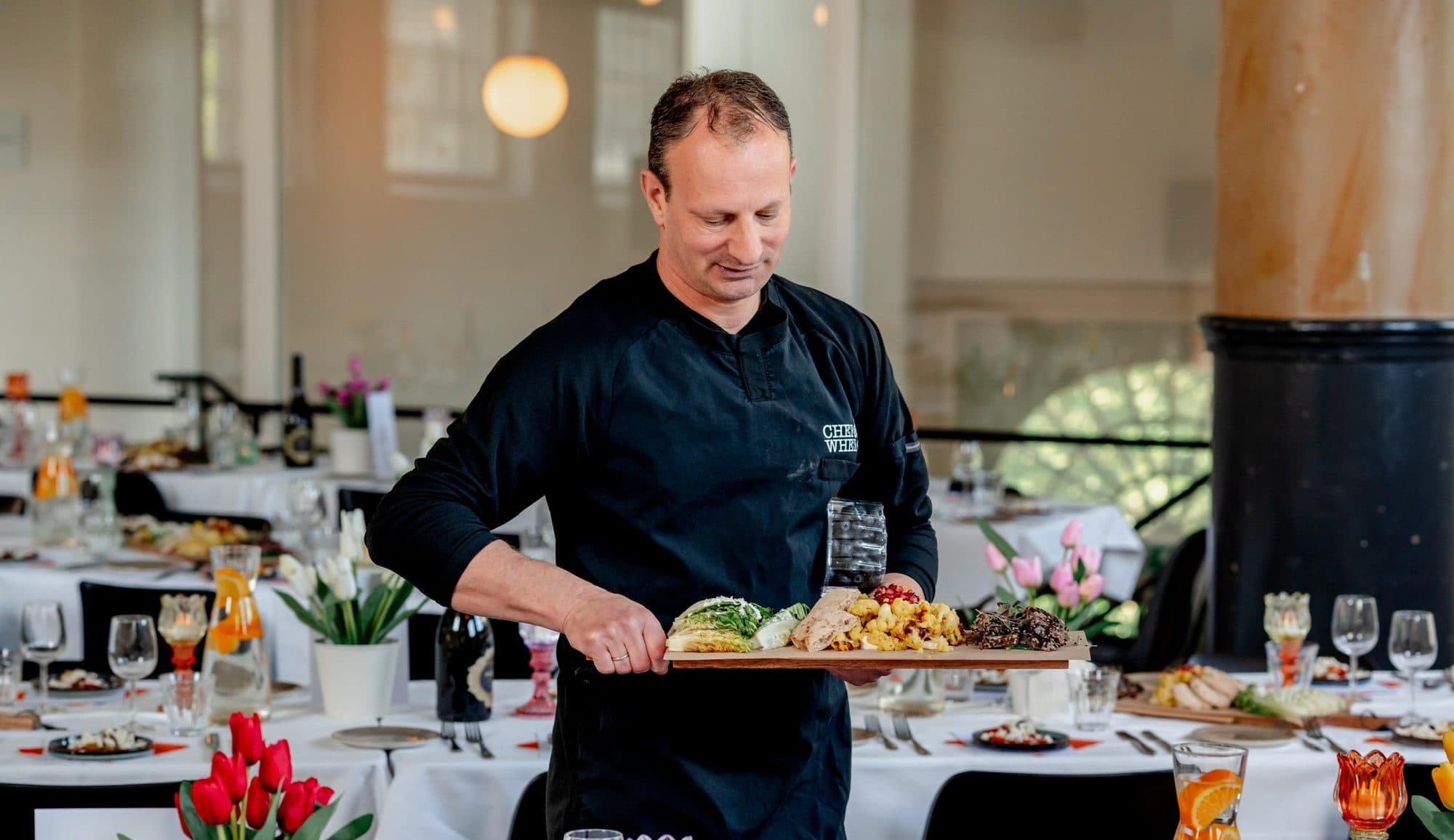 Chef Chanan loopt met een plank vol gerechten door een feestelijk gedekte zaal