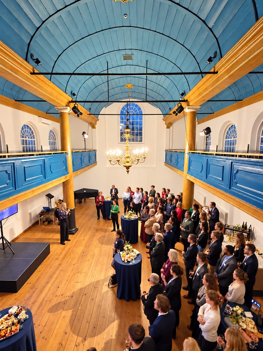 Een zakelijke bijeenkomst in een historische zaal met blauw gewelf, met een spreker en staande gasten